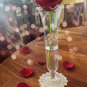 May include: A clear glass vase holding a red rose, with scattered red rose petals on a wooden table. The vase sits on a white lace doily. The image has a romantic theme, suitable for Valentine's Day.