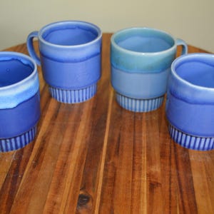 Può includere: Quattro tazze blu in ceramica con manici sono disposte su una superficie di legno. Le tazze hanno un design bicolore, con un bordo blu più chiaro e un corpo blu più scuro. La base di ogni tazza presenta delle scanalature verticali.