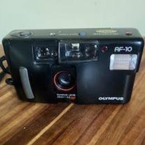 Op de afbeelding: Zwarte Olympus AF-10 35mm filmcamera met ingebouwde flitser en lens. De camera heeft een rode lensring en het Olympus-logo is zichtbaar. Op een houten oppervlak.
