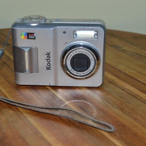 以下が含まれることがあります： 内蔵フラッシュとレンズを備えた、シルバーのKodakデジタルカメラ。カメラにはリストストラップが付いており、木製の表面に置かれています。Kodakのロゴが前面に表示されています。