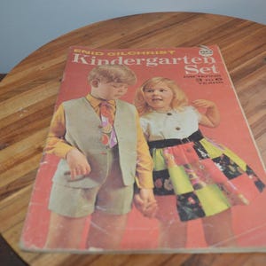 May include: A vintage Enid Gilchrist Kindergarten Set sewing pattern booklet for ages 3 to 6 years. The cover features a colour photograph of two children wearing handmade clothing, including a waistcoat, shorts, and a patchwork skirt with a white top.