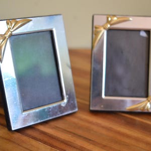 May include: Two silver picture frames with gold bow details on the corners. The square frames have a dark interior and are displayed on a wooden surface. These frames are designed to hold photographs or artwork.
