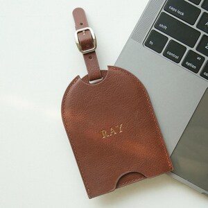 May include: A brown leather luggage tag with the name "RAY" embossed in gold lettering. The tag has a buckle closure and a strap. The tag is resting on a laptop.