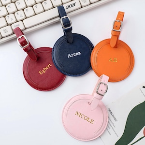 Puede incluir: Cuatro etiquetas de equipaje de cuero de colores con hebillas plateadas. Las etiquetas son rojas, azul marino, naranja y rosa. Cada etiqueta tiene un nombre o iniciales en letras doradas: "Egbert", "Aruna", "A.E." y "Nicole".