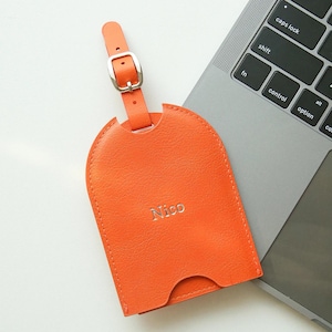 May include: An orange leather luggage tag with a silver buckle and the name "Nico" embossed on the front. The tag is arched at the top and has a slot at the bottom. The tag is lying on a laptop keyboard.