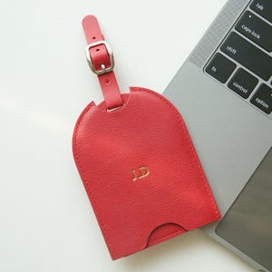 May include: A red leather luggage tag with a silver buckle and strap. The tag has a curved top and a cutout at the bottom. Gold initials "JD" are embossed on the front. The tag is next to a laptop.