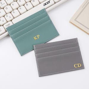 Può includere: Sono esposti due porta carte in pelle, uno verde acqua e uno grigio. Il porta carte verde acqua ha le iniziali dorate "KP", e quello grigio "CD". Entrambi hanno più fessure per carte e cuciture bianche. Una tastiera bianca è sullo sfondo.