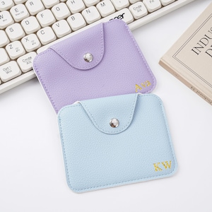 May include: Two small, square, pastel-colored pouches with flap closures and snap buttons. One is lavender, the other is light blue. Each has gold embroidered initials. A white keyboard and a book are in the background.
