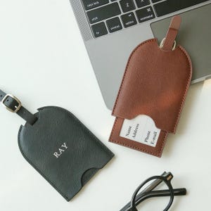 May include: Two leather luggage tags, one black with "RAY" and one brown, sit near a laptop. The brown tag has a card with "Name, Address, Phone, E-mail" visible. A pair of black glasses rests nearby.