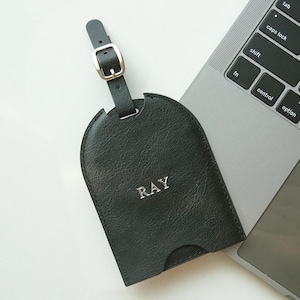 May include: A black leather luggage tag with the name "RAY" embossed in silver. The tag has a buckle closure and is next to a laptop keyboard.