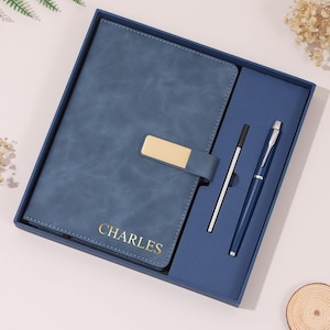 May include: A blue notebook with a gold-colored metal clasp and the name CHARLES embossed in gold on the cover. The set includes a matching blue ballpoint pen and a silver-colored pen refill, all presented in a blue gift box.