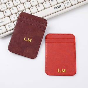 Op de afbeelding: Twee leren kaarthouders, één bordeauxrood en één rood, worden getoond. Elke kaarthouder heeft de initialen "L.M" in gouden letters. De kaarthouders zijn rechthoekig met afgeronde hoeken en hebben een vak aan de voorkant.