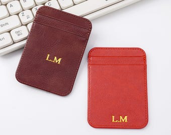 May include: Two leather cardholders, one burgundy and one red, are displayed. Each cardholder has the initials "L.M" in gold lettering. The cardholders are rectangular with rounded corners and feature a pocket on the front.