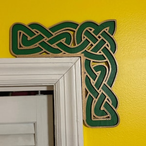 May include: A green and gold Celtic knot corner design, made of wood, is mounted on a yellow wall next to a white door frame.