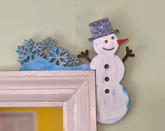 May include: A blue and white wooden snowman ornament with a blue glitter hat and a red nose. The snowman is holding up its arms and is decorated with white buttons. The snowman is placed on a white door frame with a blue snowflake design.