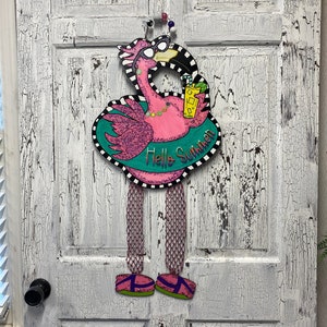 May include: A pink flamingo with sunglasses and a drink in its hand is holding a sign that says "Hello Summer". The flamingo is wearing a green and white striped swimsuit and has pink and white striped legs. The flamingo is hanging on a white door.