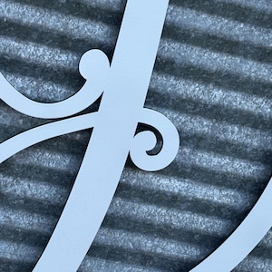 K&ouml;nnte beinhalten: Ein mattes, hellgraues Wandkunstobjekt aus Metall, das einen gro&szlig;en, eleganten, kursiven Buchstaben J darstellt. Das Design umfasst dekorative Schn&ouml;rkel-Akzente, die sich vom vertikalen Hauptstrich des Buchstabens nach au&szlig;en biegen.