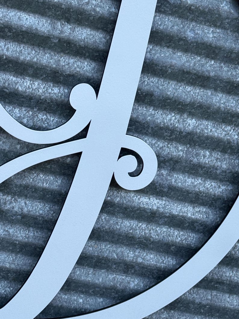 K&ouml;nnte beinhalten: Ein mattes, hellgraues Wandkunstobjekt aus Metall, das einen gro&szlig;en, eleganten, kursiven Buchstaben J darstellt. Das Design umfasst dekorative Schn&ouml;rkel-Akzente, die sich vom vertikalen Hauptstrich des Buchstabens nach au&szlig;en biegen.