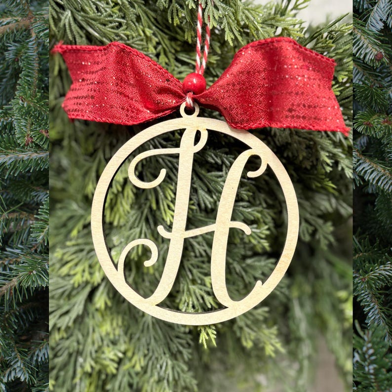 K&ouml;nnte beinhalten: Ein kreisf&ouml;rmiges Ornament aus hellem Holz mit einem gro&szlig;en, stilisierten Buchstaben H als Ausschnitt in der Mitte. Das Ornament ist mit einer leuchtend roten Stoffschleife, einer kleinen roten Perle und einer rot-wei&szlig;en Kordel versehen.