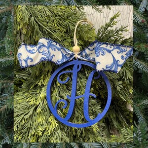 K&ouml;nnte beinhalten: Ein kreisf&ouml;rmiges blaues Holzornament mit einem ausgeschnittenen Buchstaben H in der Mitte. Das Ornament h&auml;ngt an einer hellen Schnur mit einer kleinen Holzperle und ist mit einem wei&szlig;en Stoffband mit blauem Wirbel-Blumenmuster verziert.