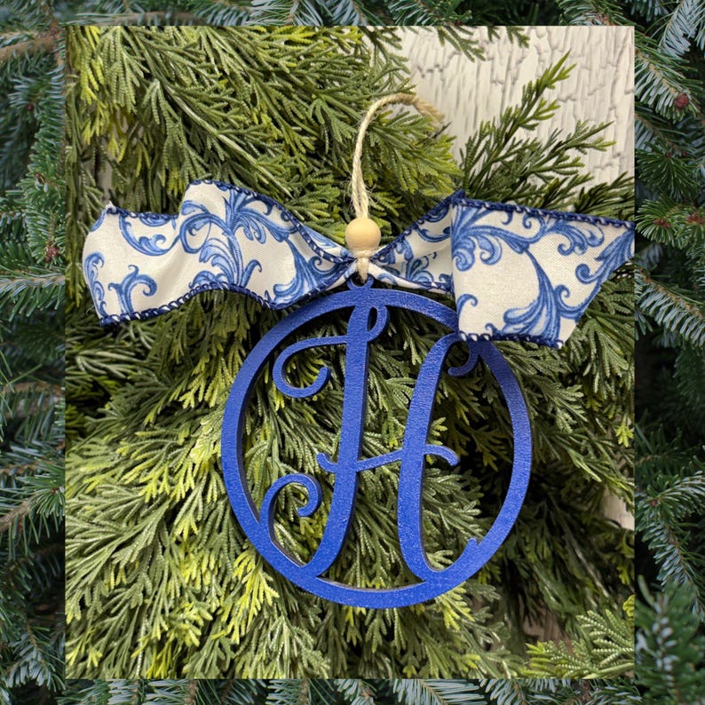 K&ouml;nnte beinhalten: Ein kreisf&ouml;rmiges blaues Holzornament mit einem ausgeschnittenen Buchstaben H in der Mitte. Das Ornament h&auml;ngt an einer hellen Schnur mit einer kleinen Holzperle und ist mit einem wei&szlig;en Stoffband mit blauem Wirbel-Blumenmuster verziert.