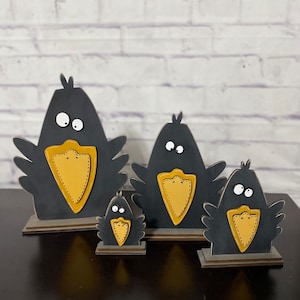 May include: A set of four black wooden crow figurines with yellow beaks and white eyes. The crows are standing on wooden bases and are arranged in order of size, with the largest crow in the back and the smallest crow in the front.