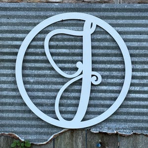 K&ouml;nnte beinhalten: Ein kreisf&ouml;rmiges Wandschild mit einem gro&szlig;en, stilisierten, kursiven Buchstaben J in mattwei&szlig;er Ausf&uuml;hrung. Der Buchstabe ist in einem d&uuml;nnen, passenden wei&szlig;en kreisf&ouml;rmigen Rahmen zentriert, was ein klares, monochromes Design ergibt.