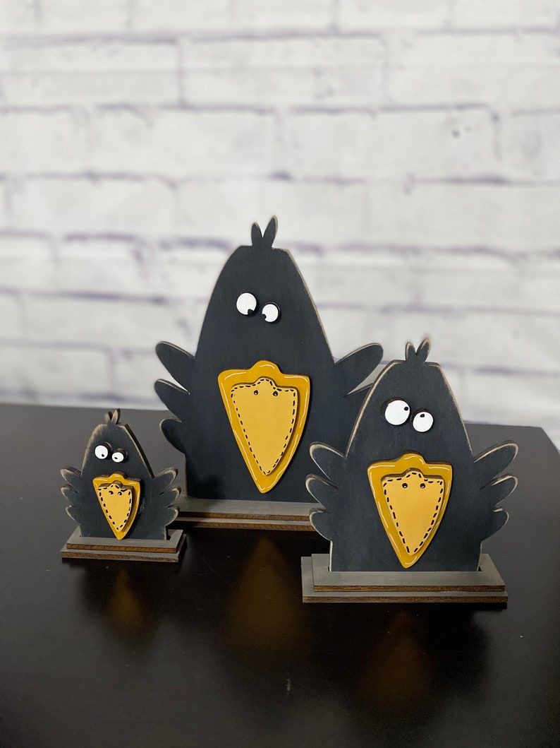 May include: Three black wooden crow figurines with yellow beaks and white eyes. The crows are different sizes and are standing on wooden bases.