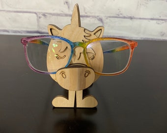 May include: Wooden unicorn-shaped eyeglasses holder with a rainbow-colored frame. The unicorn has a horn, closed eyes, and a small mouth. The holder is standing on a dark surface.