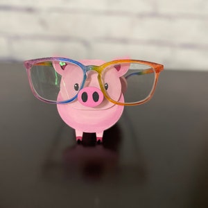 May include: A pink pig figurine wearing rainbow-colored glasses. The pig has a small snout and two black eyes.
