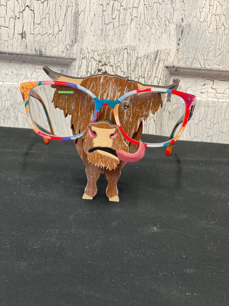 May include: A wooden Highland Cow figurine wearing colorful glasses. The cow has a pink tongue sticking out. The figurine is a fun and quirky way to display glasses.