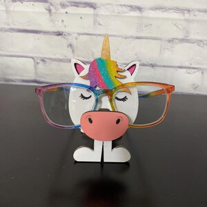 May include: A white unicorn-shaped glasses holder with a rainbow mane and a pink nose. The unicorn is wearing glasses.