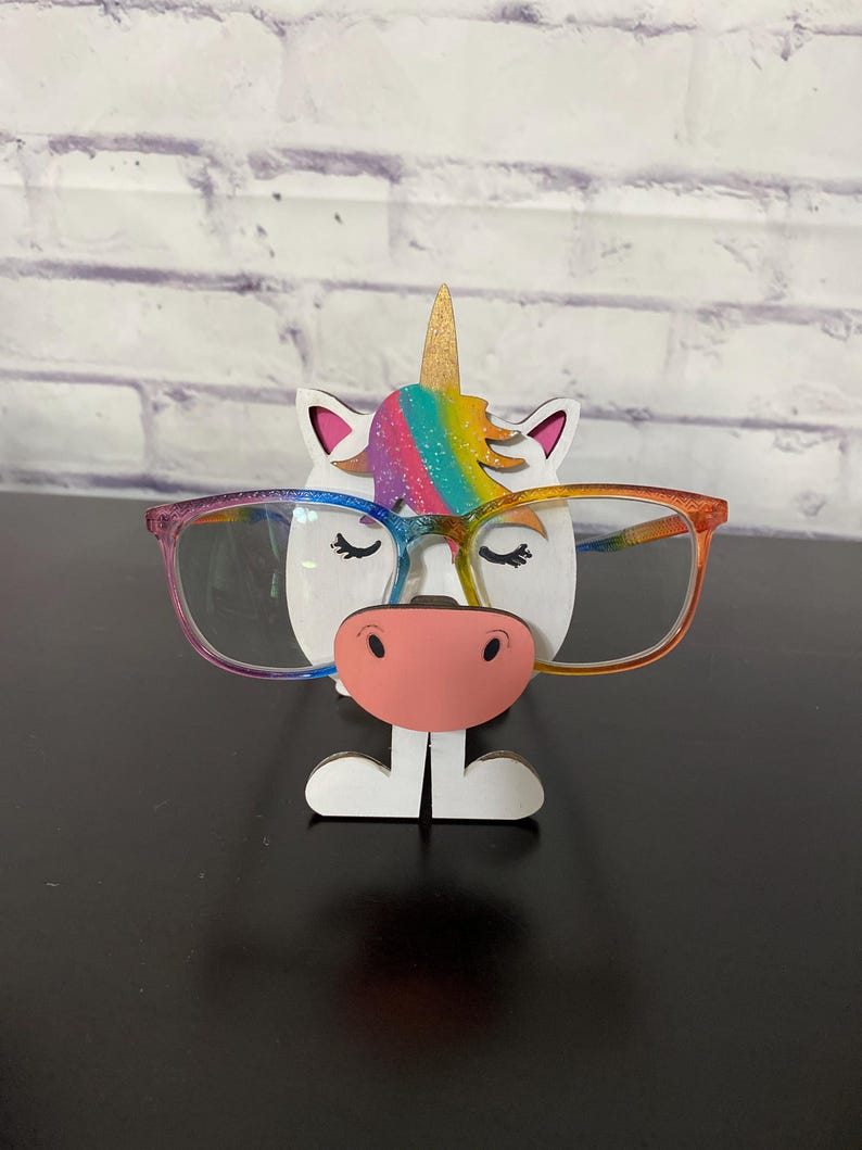 May include: A white unicorn-shaped glasses holder with a rainbow mane and a pink nose. The unicorn is wearing glasses.