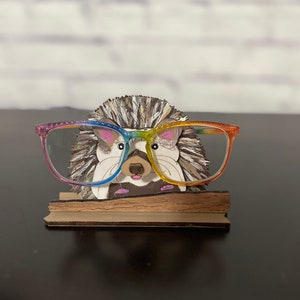May include: A wooden hedgehog figurine wearing rainbow-colored glasses, holding a pair of glasses on a wooden stand.