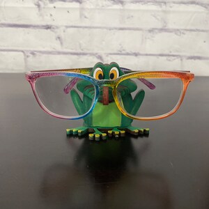 May include: A green frog-shaped glasses holder with rainbow-colored accents. The holder is holding a pair of clear glasses with rainbow-colored frames.