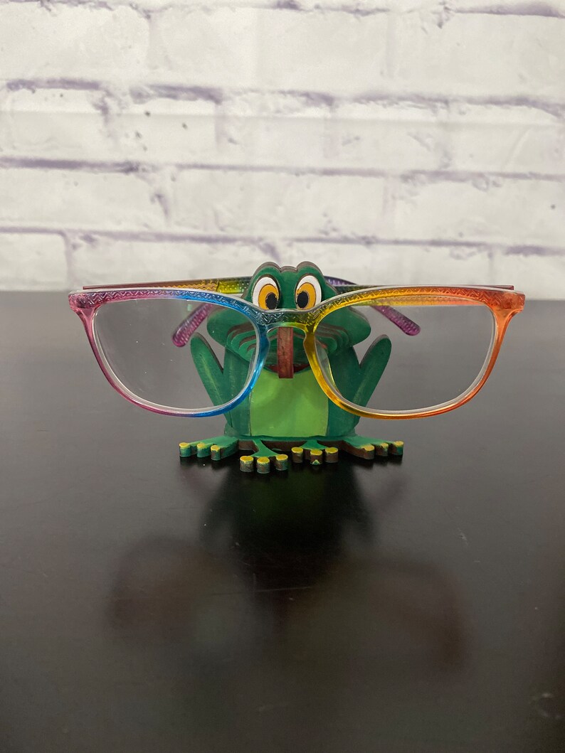 May include: A green frog-shaped glasses holder with rainbow-colored accents. The holder is holding a pair of clear glasses with rainbow-colored frames.