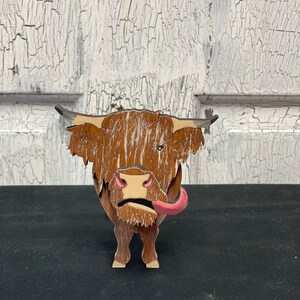 May include: A wooden figurine of a Highland cow with brown fur, a pink tongue sticking out, and a white background.
