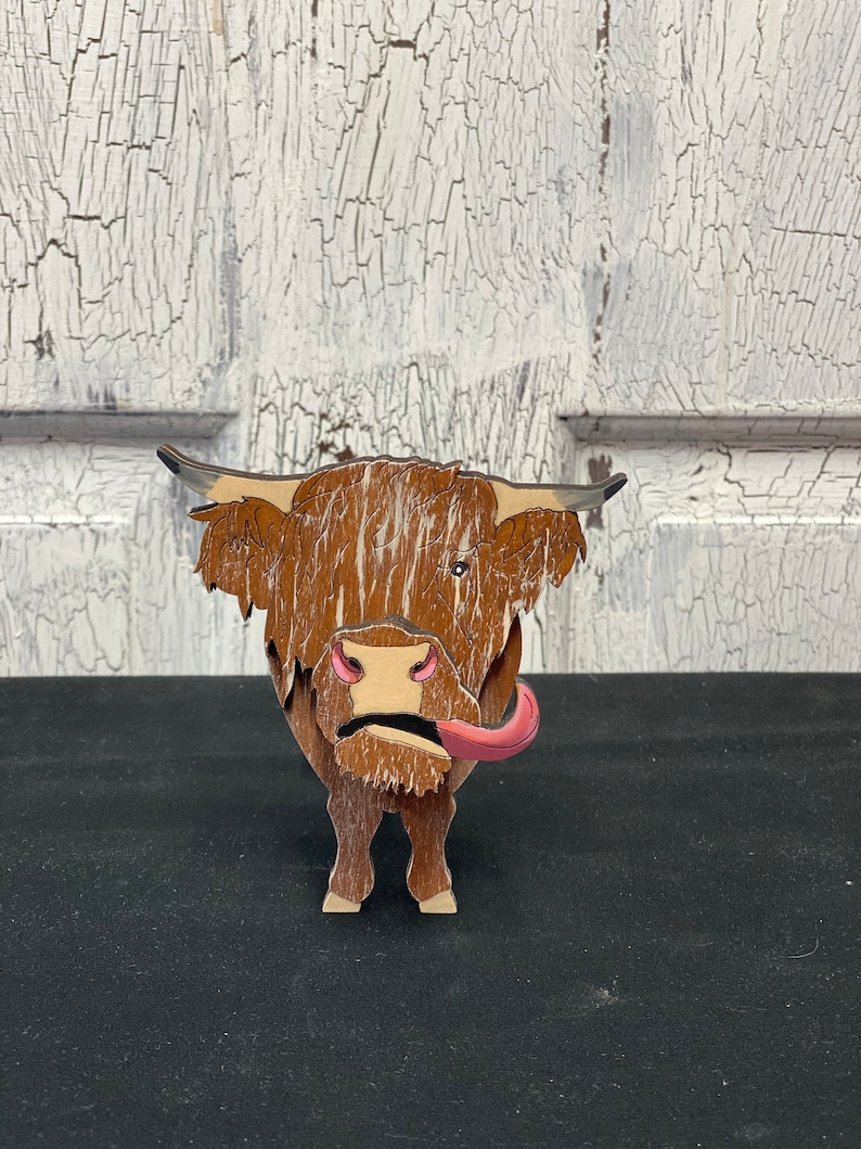 May include: A wooden figurine of a Highland cow with brown fur, a pink tongue sticking out, and a white background.