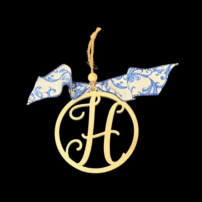 K&ouml;nnte beinhalten: Ein kreisf&ouml;rmiges Ornament aus hellem Holz mit einem ausgeschnittenen Buchstaben H in der Mitte. Das Ornament ist an einer beigen Schnurschlaufe mit einer kleinen Holzperle aufgeh&auml;ngt und mit einer blau-wei&szlig; gemusterten Stoffschleife verziert.
