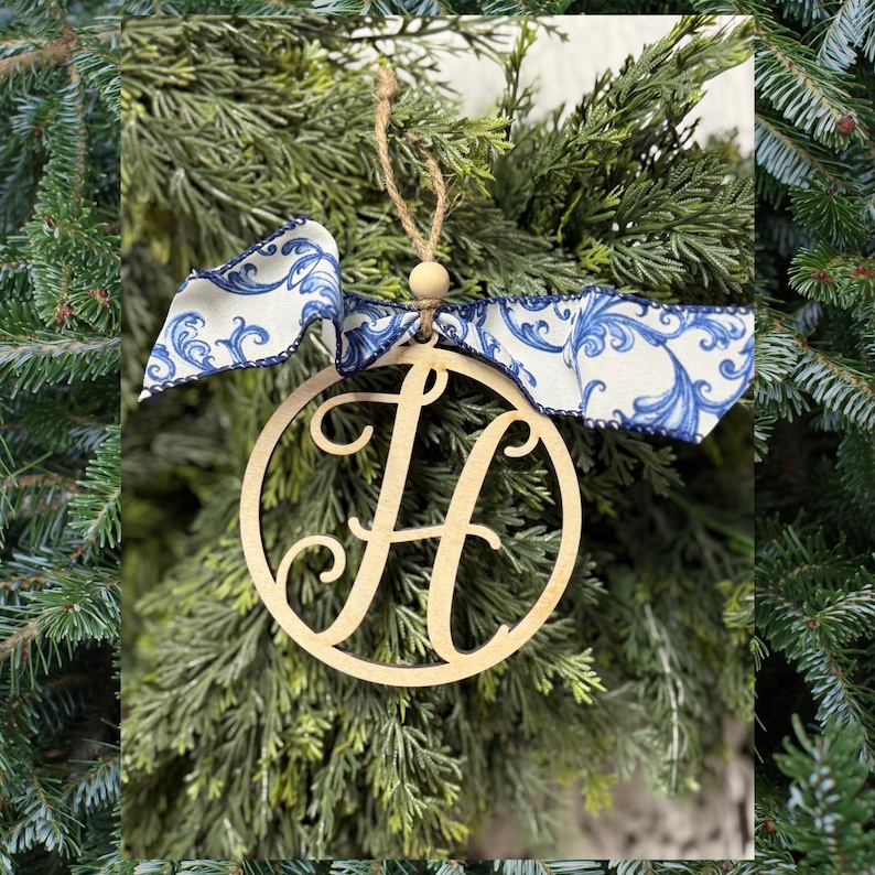 K&ouml;nnte beinhalten: Ein kreisf&ouml;rmiges Holzornament mit einem ausgeschnittenen Buchstaben H in Schreibschrift. Das Ornament ist mit einem dekorativen wei&szlig;en Band mit blauem Blumenmuster versehen und an einer d&uuml;nnen Juteschnur mit einer kleinen Holzperle befestigt.
