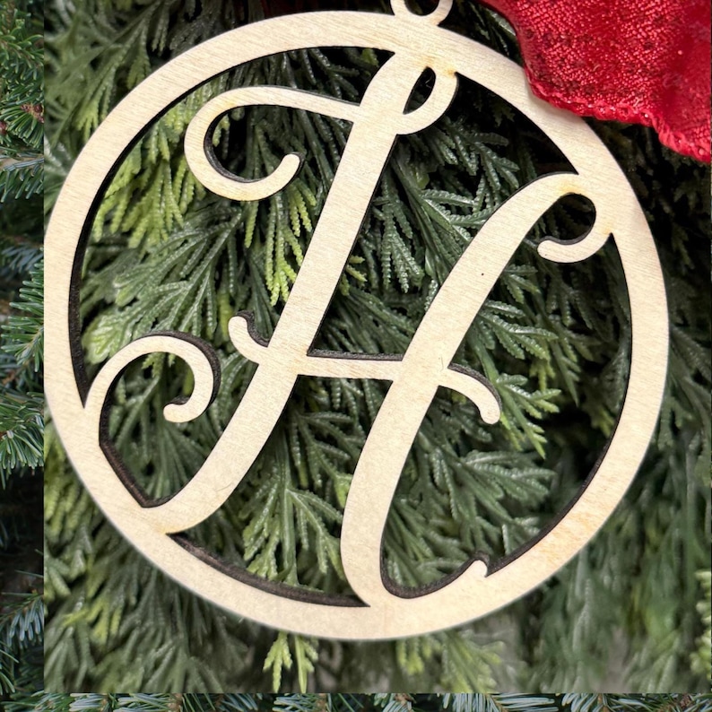 K&ouml;nnte beinhalten: Ein kreisf&ouml;rmiges Ornament aus hellem Holz mit einem gro&szlig;en, ausgeschnittenen Buchstaben H in Schreibschrift in der Mitte. Das Ornament hat eine glatte Naturholzoberfl&auml;che mit dunklen, lasergeschnittenen Kanten und ein rotes Stoffband.