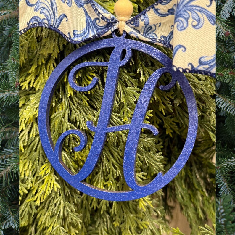 K&ouml;nnte beinhalten: Ein kreisf&ouml;rmiges blaues Holzornament mit einem gro&szlig;en stilisierten Buchstaben H in der Mitte. Das Ornament ist an einem cremefarbenen Band mit blauem Blumenmuster und einer kleinen Holzperle an der Oberseite befestigt, ein dekoratives St&uuml;ck.