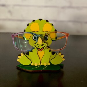 May include: A green dinosaur-shaped glasses holder with a yellow belly and black spots. The dinosaur is sitting on a green leaf and wearing rainbow-colored glasses.