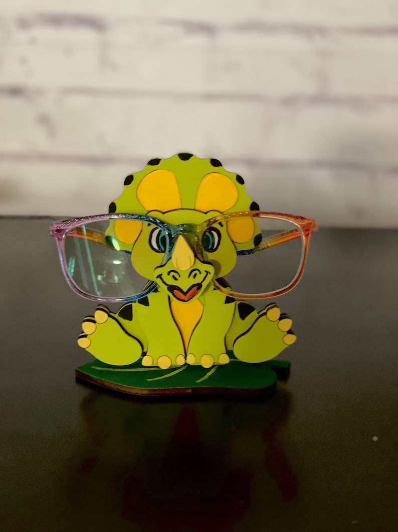 May include: A green dinosaur-shaped glasses holder with a yellow belly and black spots. The dinosaur is sitting on a green leaf and wearing rainbow-colored glasses.