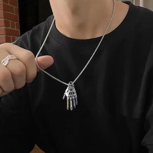 May include: Silver-coloured necklace with a pendant in the shape of a skeletal hand, with gold-coloured fingertips. The pendant hangs from a fine chain. The person is wearing a black top.