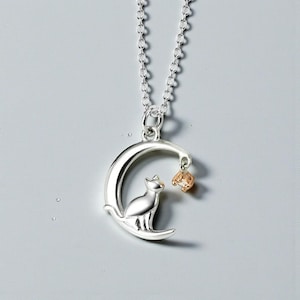 May include: Silver-toned cat pendant necklace. The pendant features a cat silhouette seated on a crescent moon, with a small, gold-colored charm suspended from the moon's tip. The necklace chain is also silver.