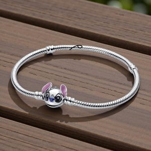 May include: A silver bracelet featuring a charm of the character Stitch from the Disney movie Lilo & Stitch. The charm is white with pink ears and blue accents. The bracelet is on a wooden surface.