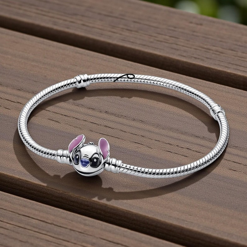 May include: A silver bracelet featuring a charm of the character Stitch from the Disney movie Lilo & Stitch. The charm is white with pink ears and blue accents. The bracelet is on a wooden surface.