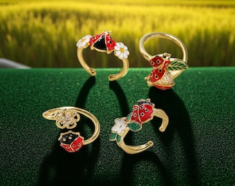 May include: Four gold-toned rings with ladybug and floral designs. The ladybugs are red and black with white accents. One ring features a ladybug with a daisy, another with a leaf, and another with a flower.