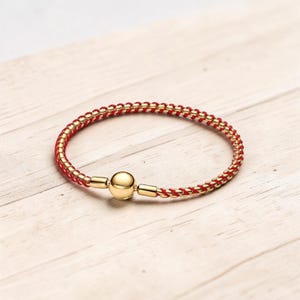 May include: A gold-colored bracelet with a red woven design. The bracelet features a gold clasp with a spherical center. The bracelet is displayed on a light-colored wooden surface.
